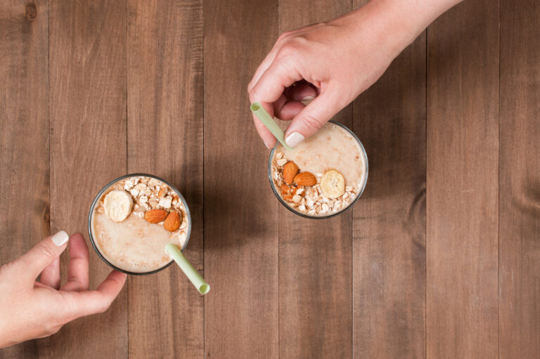 Tangan perempuan memegang gelas smoothie oatmeal pisang dengan topping irisan pisang dan oat di atas meja.
