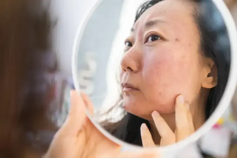 Wanita muda Asia menatap kulit wajahnya sambil menyentuh pipi, menggambarkan perawatan kulit dan kecantikan.
