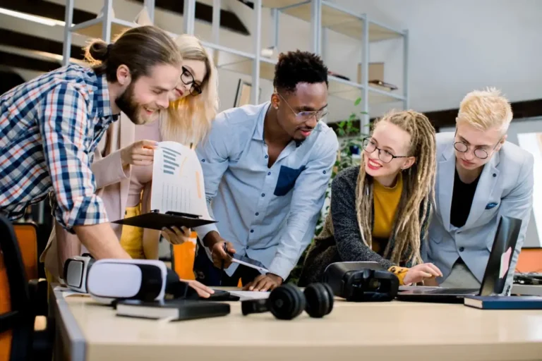 Sekelompok pebisnis muda multietnis tersenyum sambil berdiskusi di kantor modern dengan laptop dan dokumen.