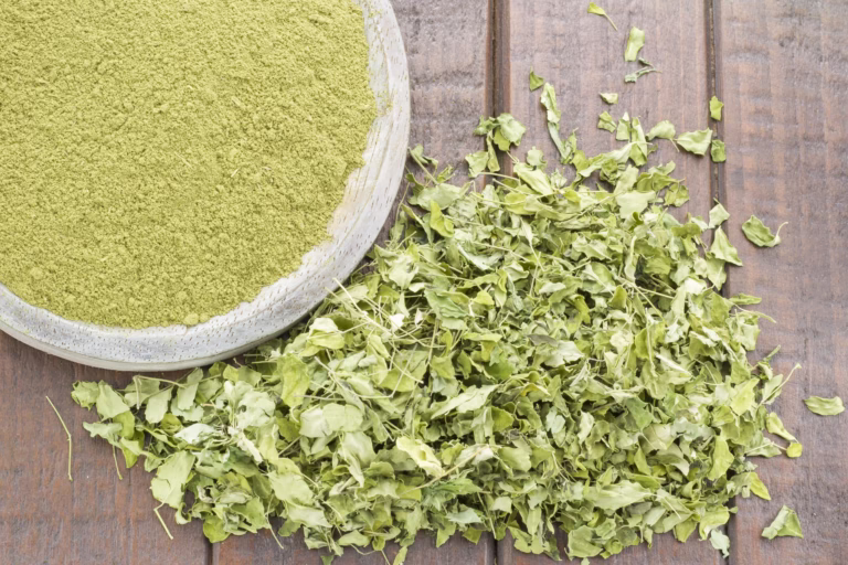 Bubuk daun kelor hijau dalam mangkuk kayu dengan daun kelor segar di meja, menonjolkan bahan herbal alami.
